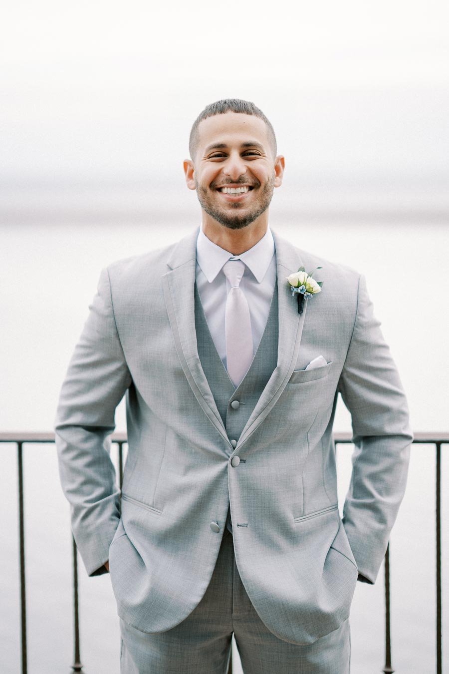 ```markdown Smiling groom in a grey suit with a boutonniere, posing outdoors by a lakeside railing on a cloudy day. ```