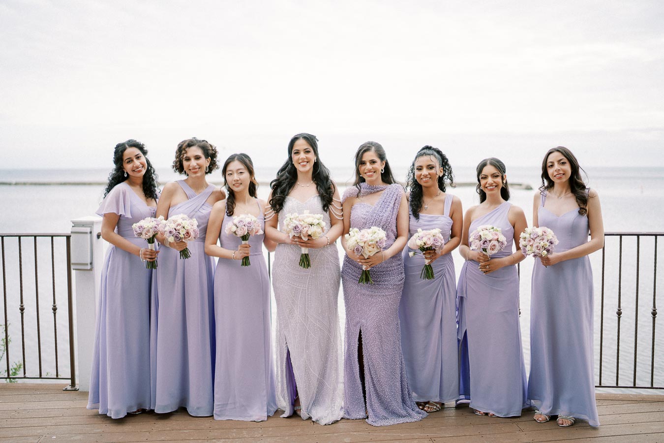 Bridesmaids in lavender dresses holding bouquets, standing together by a lakeside on a cloudy day.