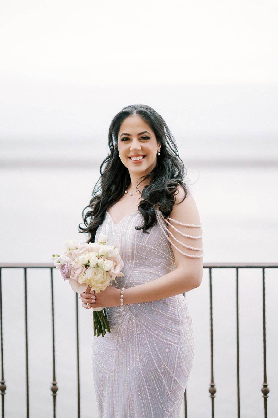 Elegant bride in a sparkling gown holding a pastel bouquet, smiling by a lakeside railing background.