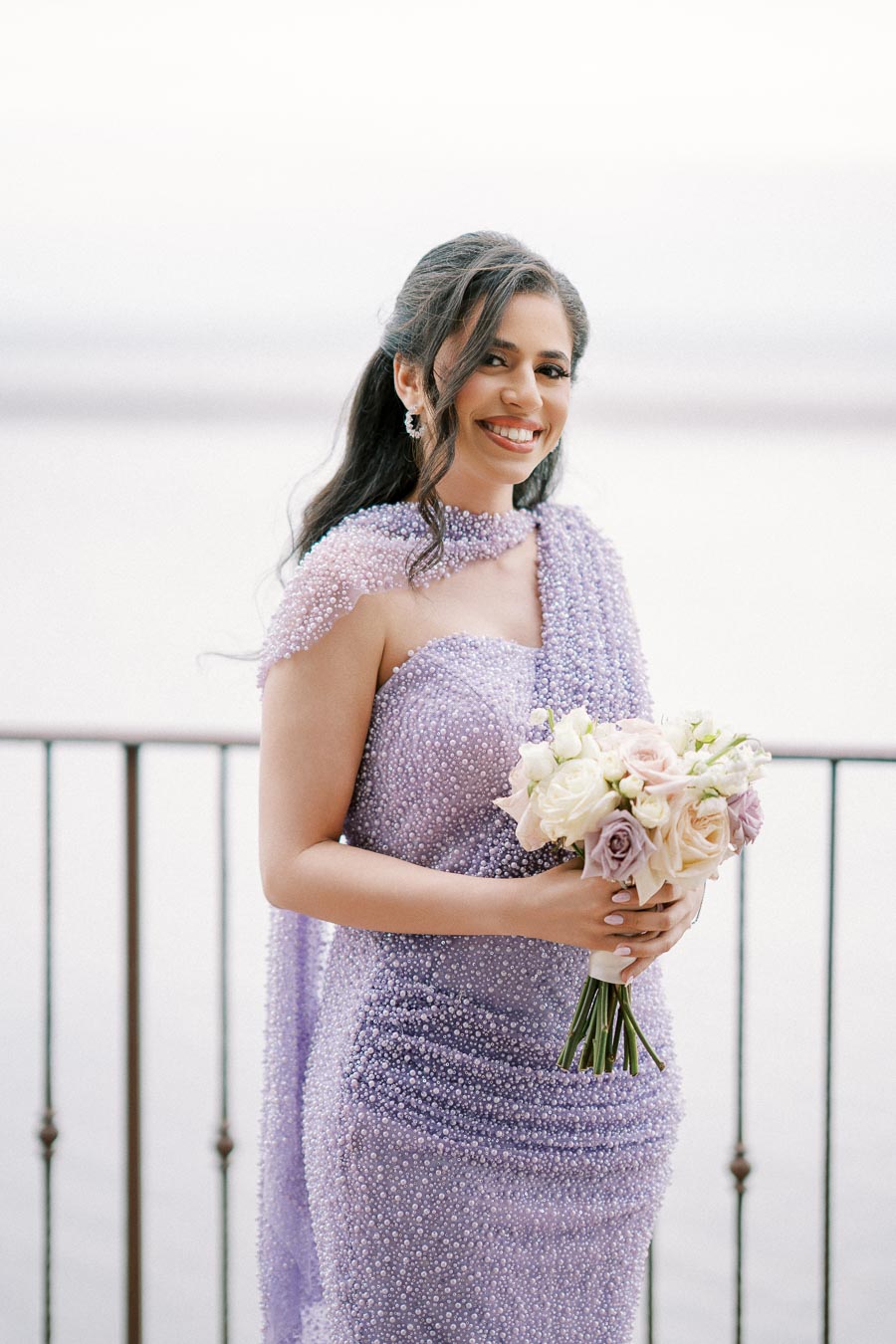 Young woman in a sparkling lilac gown holding a bouquet of pastel flowers, overlooking a scenic water view with a serene expression.