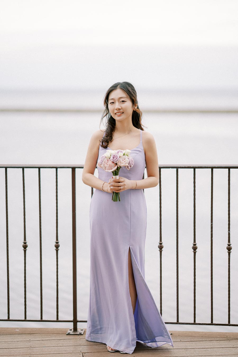 A woman in a lavender bridesmaid dress holding a bouquet of pink and white roses, standing on a waterfront balcony.