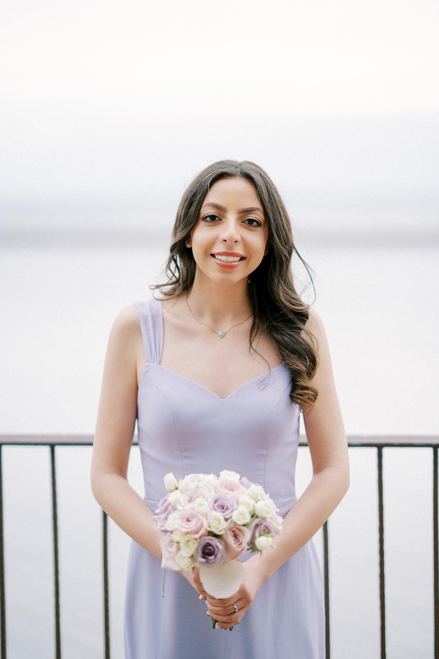 Young woman in a lavender dress holding a bouquet of pastel roses by a lakeside railing.