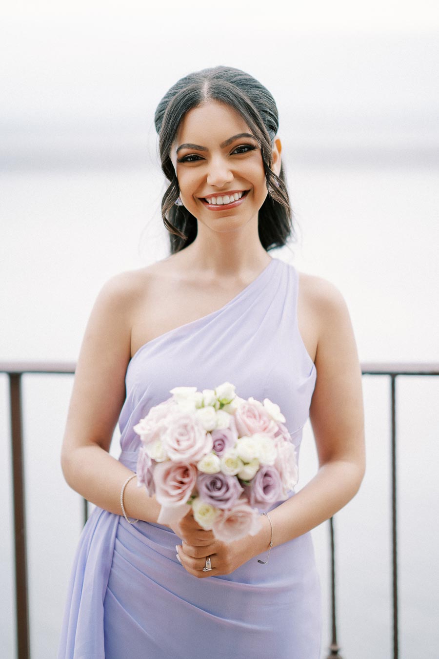 Smiling woman in lavender one-shoulder dress holding a bouquet of pink and white roses, standing outdoors with a serene backdrop.