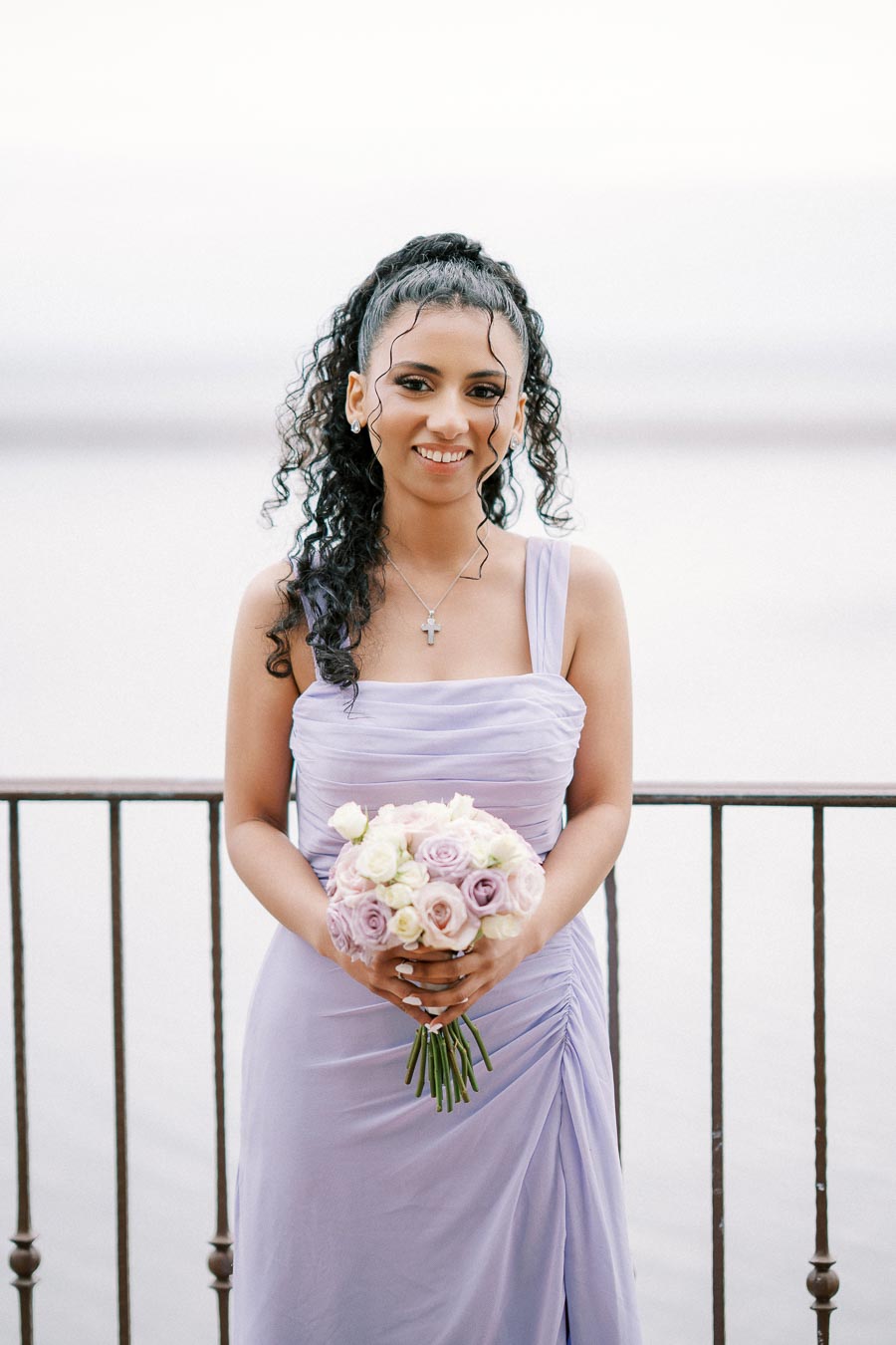 Young woman in a lavender bridesmaid dress holding a bouquet of pink and white roses, standing by a railing with a serene water backdrop.