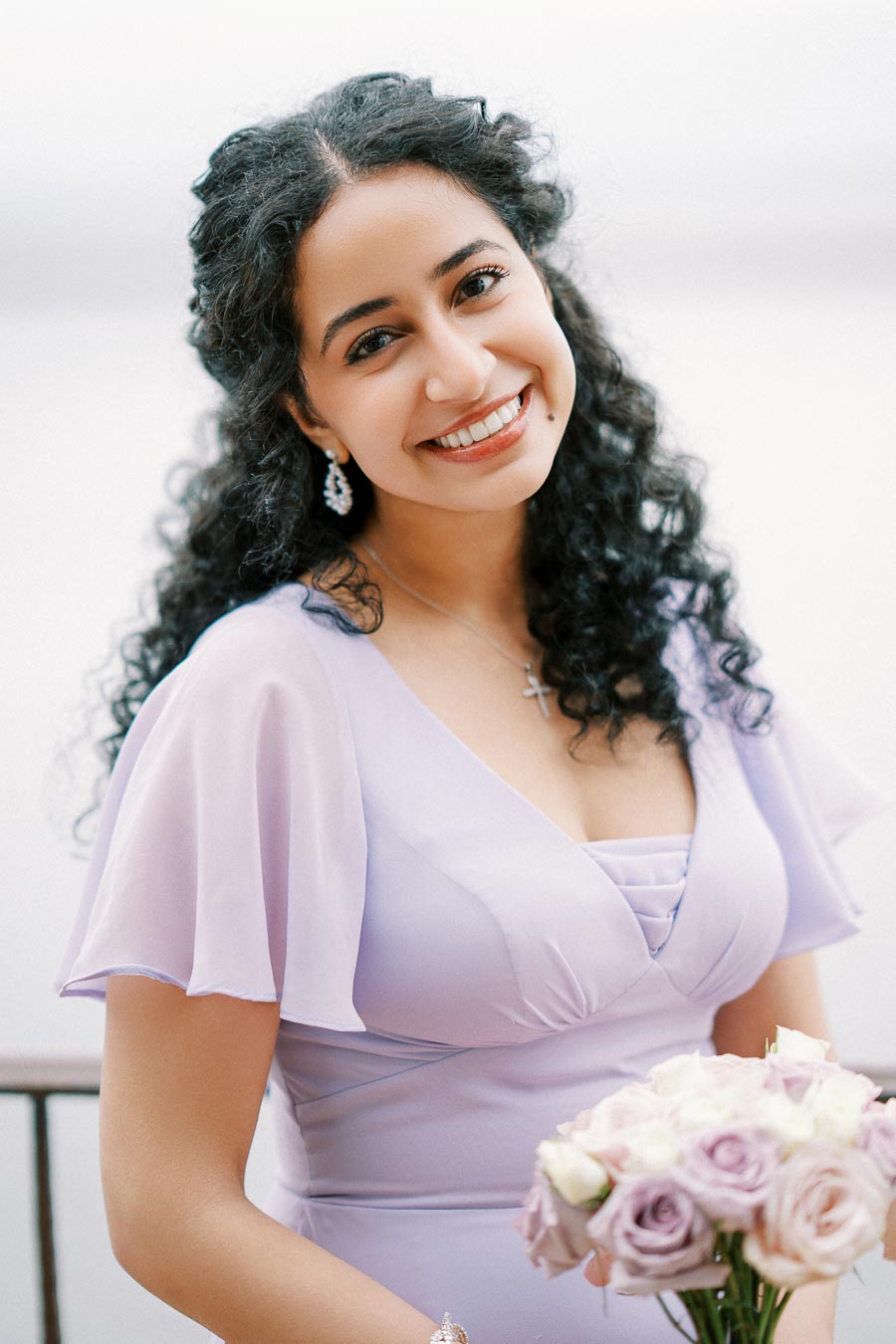 Smiling woman with curly hair holding a bouquet, wearing a light purple dress, standing outdoors.