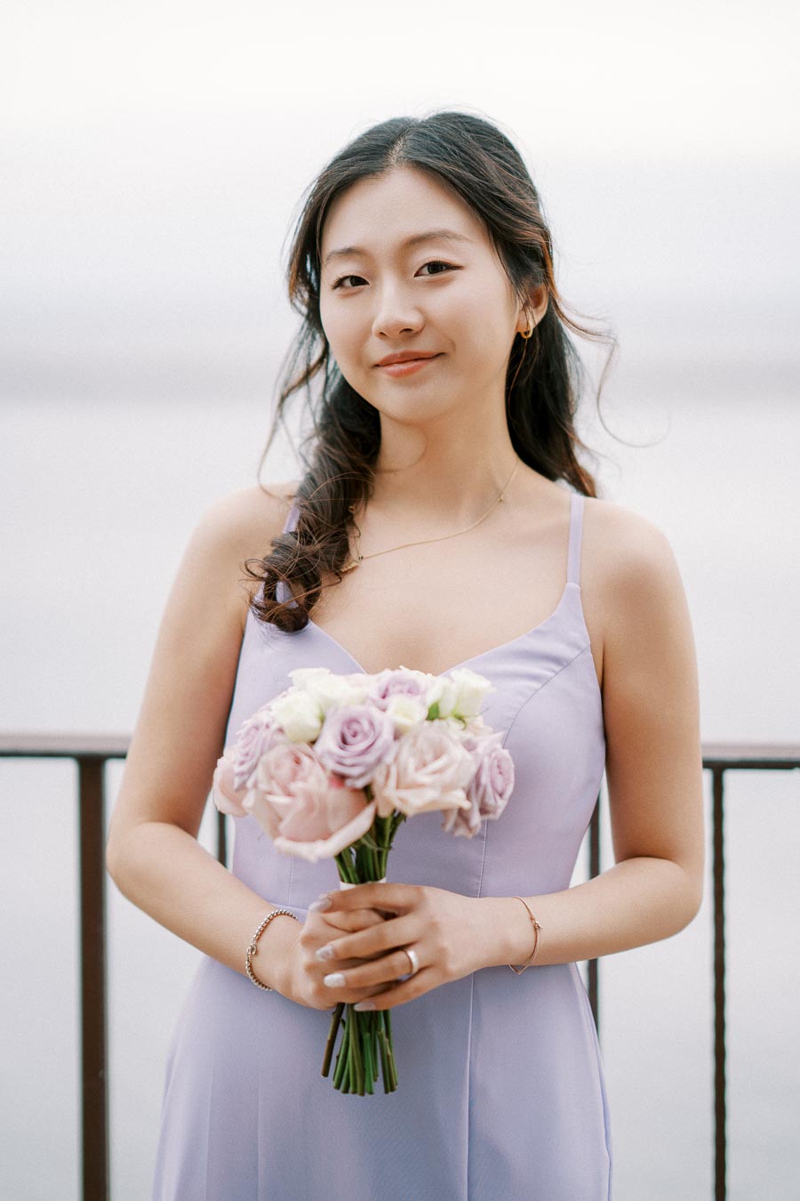 Young woman in a lavender dress holding a bouquet of pink and white roses, standing by a railing with a serene background.