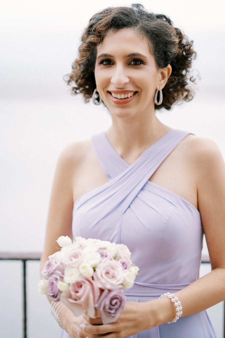 A woman in a lavender bridesmaid dress smiling and holding a bouquet of pink and white roses, standing outdoors on a wedding day.