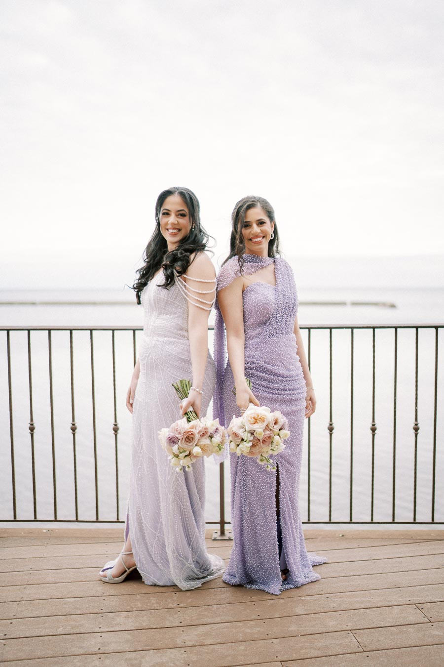 Two women in elegant lavender gowns, smiling and holding bouquets of pastel flowers, standing on a wooden deck by the water with a railing in the background.