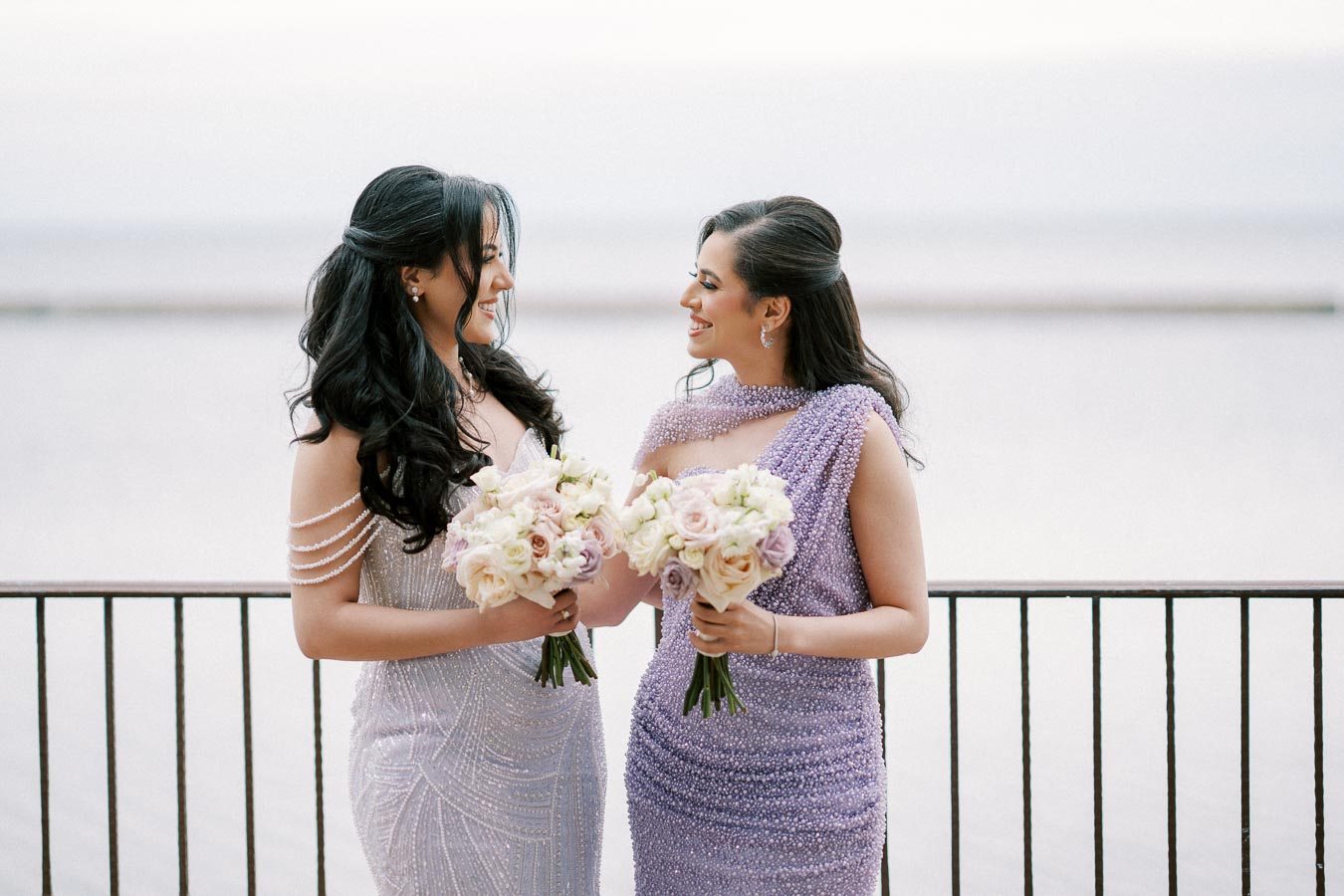 Two women in elegant, beaded gowns holding flower bouquets and smiling at each other, standing by a waterfront.
