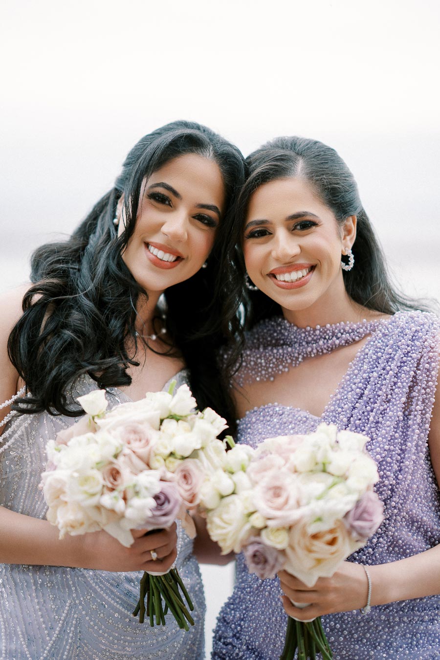 Two women smiling and holding elegant bouquets of pastel roses, dressed in beaded gowns, capturing a joyful moment at a special event.