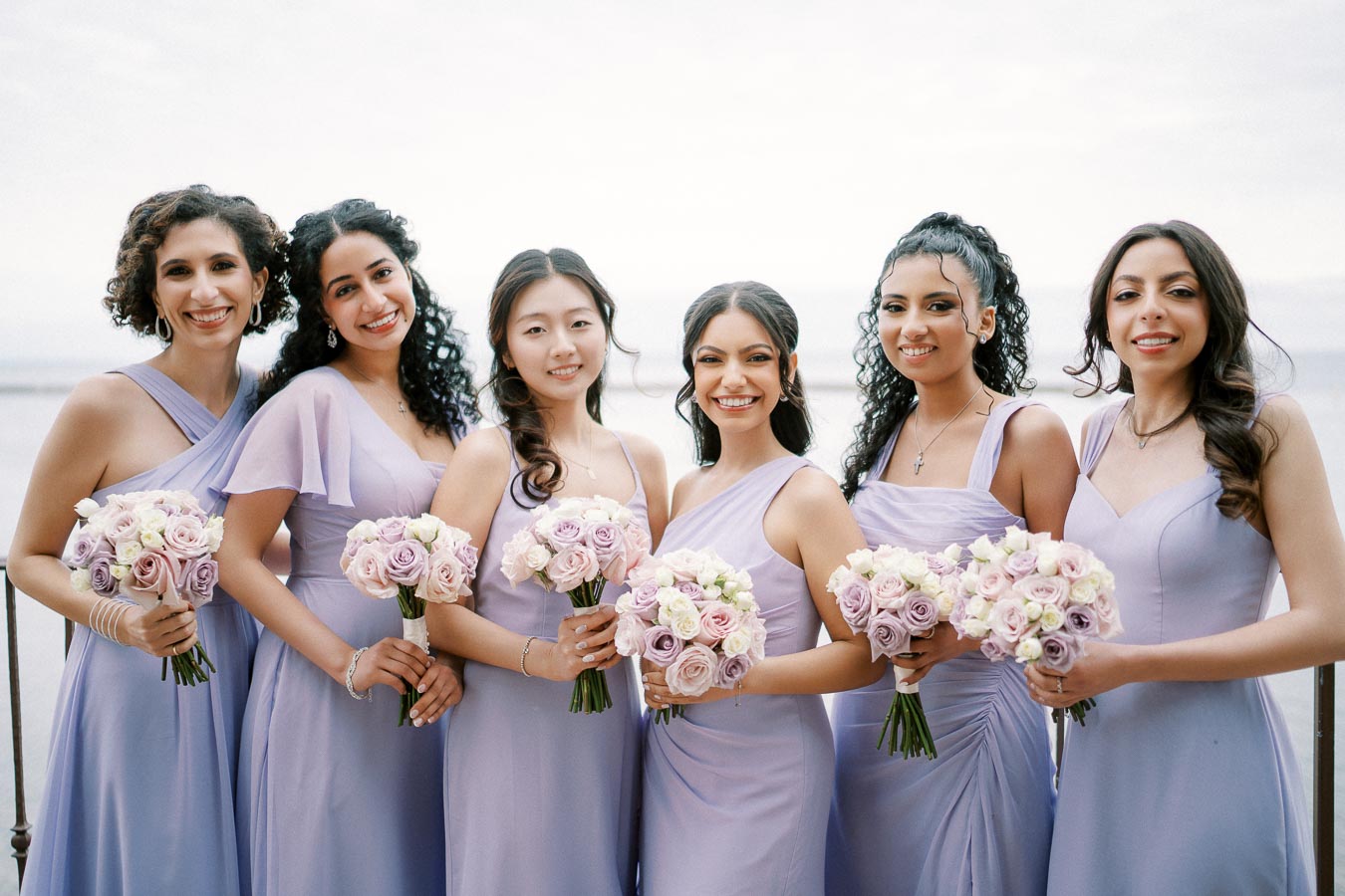A group of six bridesmaids in lavender dresses holding bouquets of roses, smiling and standing together outdoors with a scenic background.