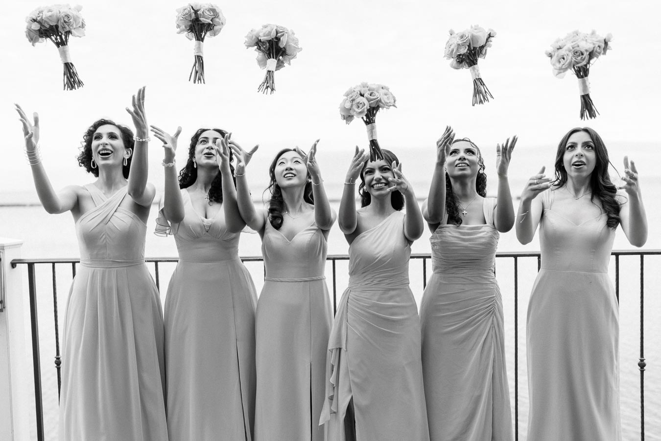 Seven bridesmaids in elegant dresses joyfully toss bouquets into the air against a scenic outdoor backdrop.