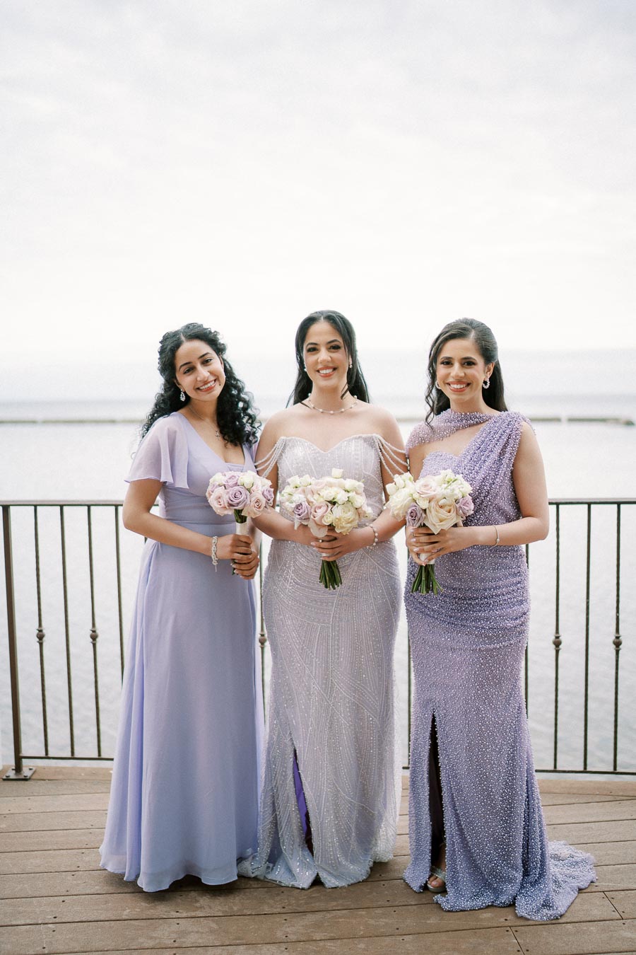Three women in elegant lavender and silver dresses holding bouquets, standing on a wooden deck by a waterfront, perfect for a bridesmaid or formal event theme.