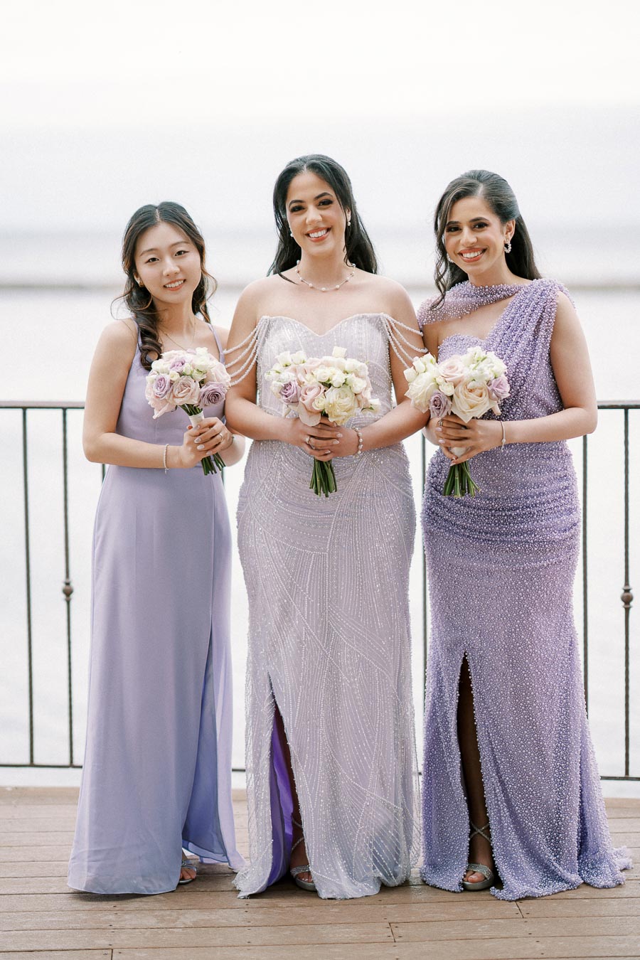 Three women in elegant lavender dresses holding floral bouquets, standing on a wooden terrace with a scenic water background.
