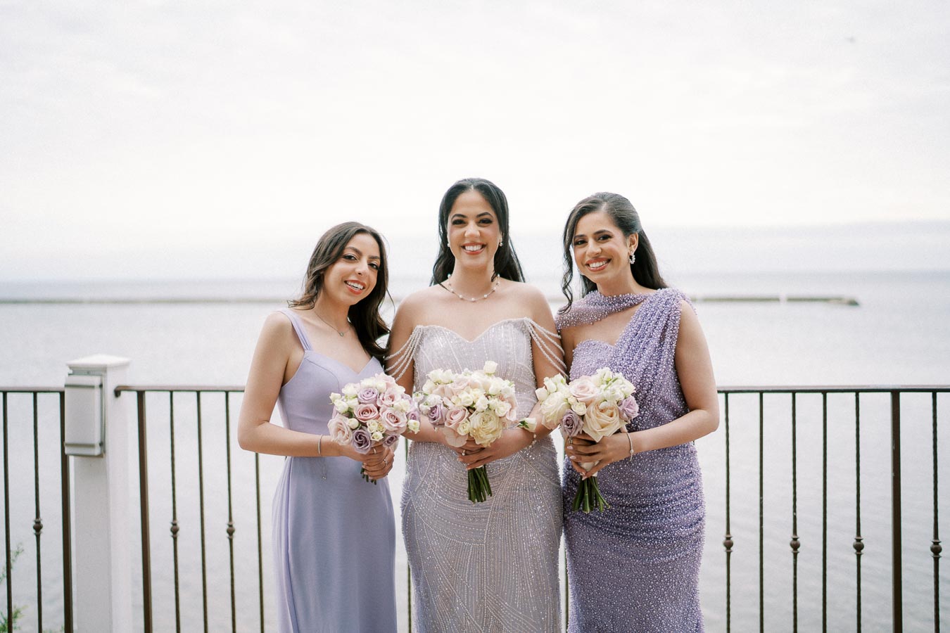 Three women in elegant evening gowns holding bouquets of pastel flowers, standing in front of a seaside backdrop with a metallic railing.