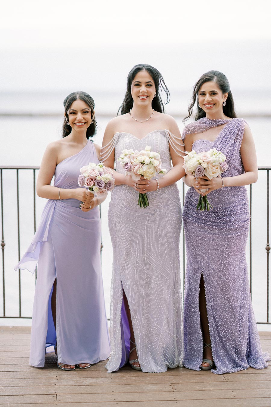 Three women in elegant lavender and lilac gowns, holding pastel bouquets, stand on a wooden deck overlooking the ocean.