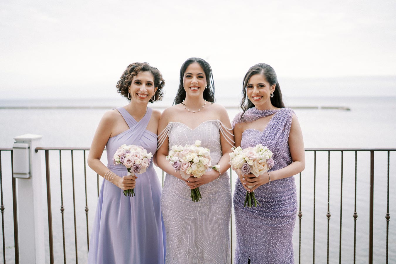 Bridesmaids in elegant lavender dresses holding bouquets, posing together on a waterfront balcony with a serene ocean view in the background.