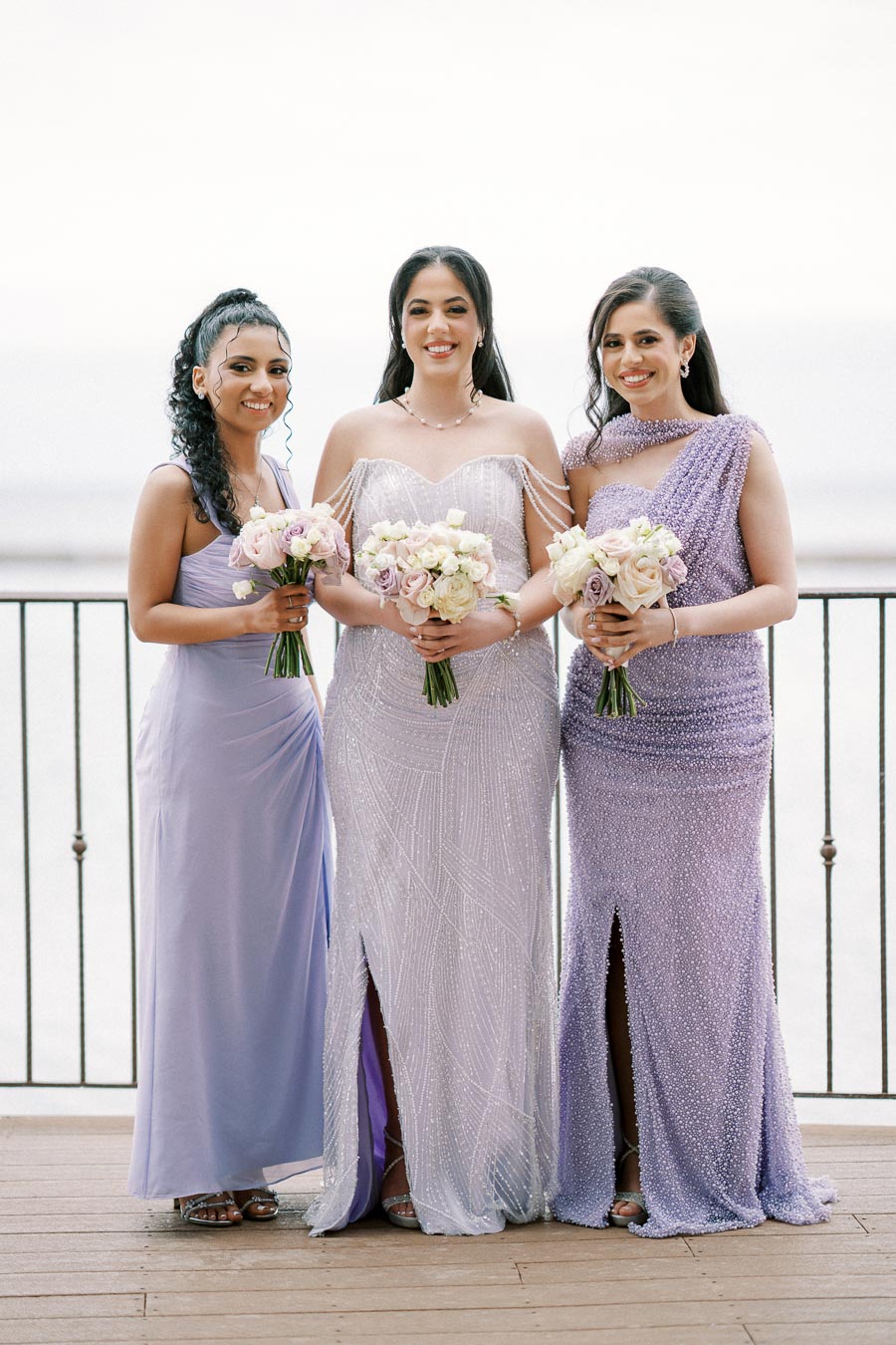 Three women in elegant lavender and lilac dresses holding bouquets of flowers, posing on a wooden deck with a scenic waterfront background.