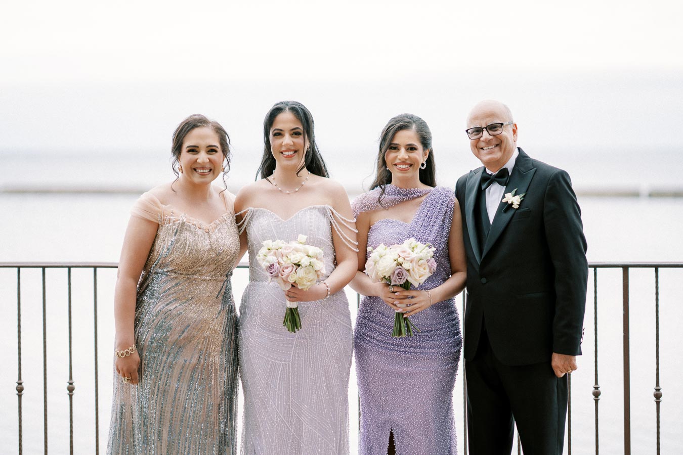 A group of people dressed elegantly at a wedding, featuring three women in beaded gowns holding flower bouquets and a man in a tuxedo, posing in front of a scenic lakeside backdrop.