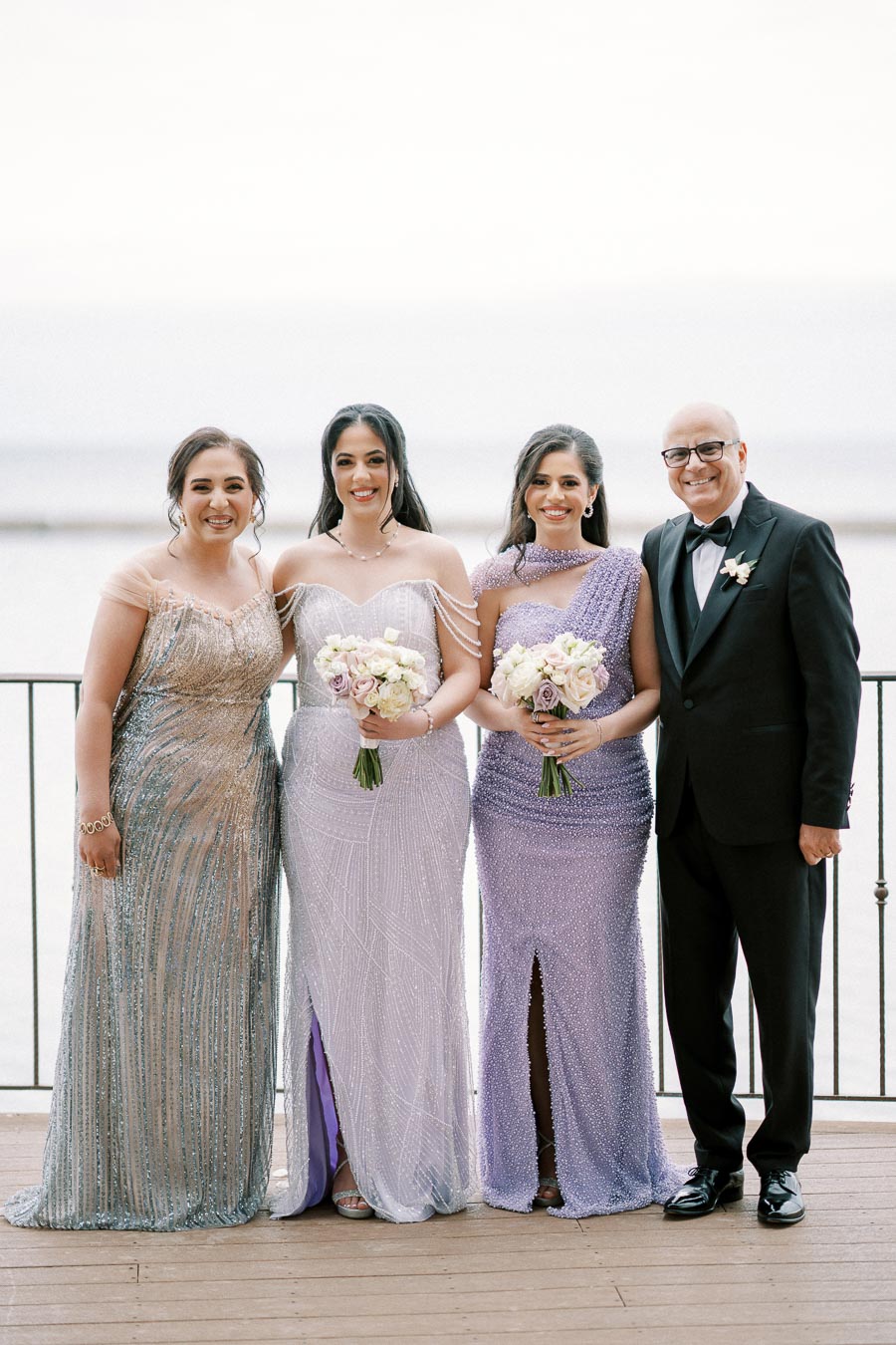 A group of elegantly dressed individuals posing together on a balcony with a scenic waterfront backdrop; women in sparkling gowns holding floral bouquets and a man in a tuxedo smiling.