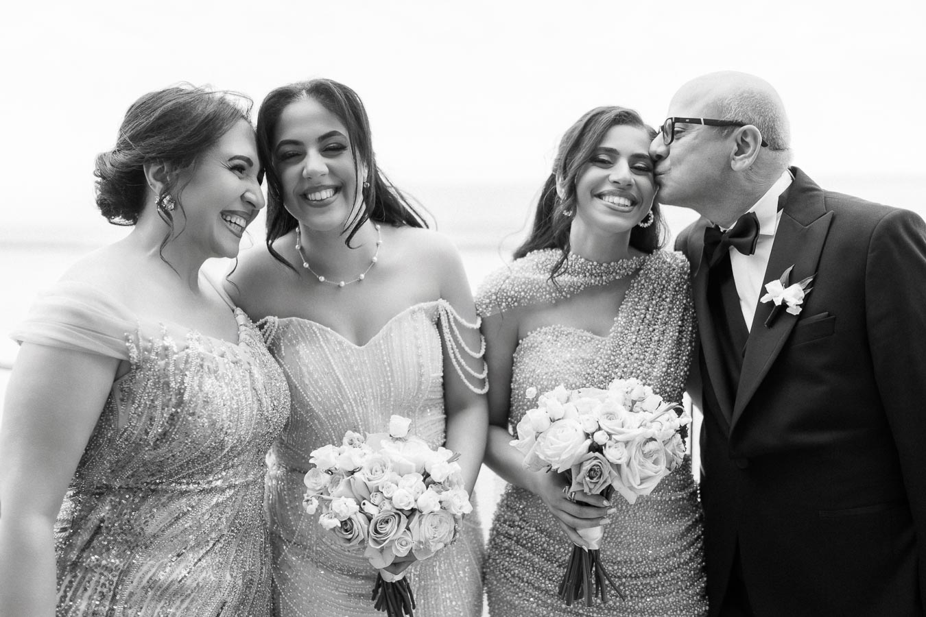 A joyful black and white wedding photo showing a family celebrating together. Two women in elegant dresses hold bouquets of flowers, smiling brightly, while a third woman receives a kiss on the cheek from a man in a tuxedo.