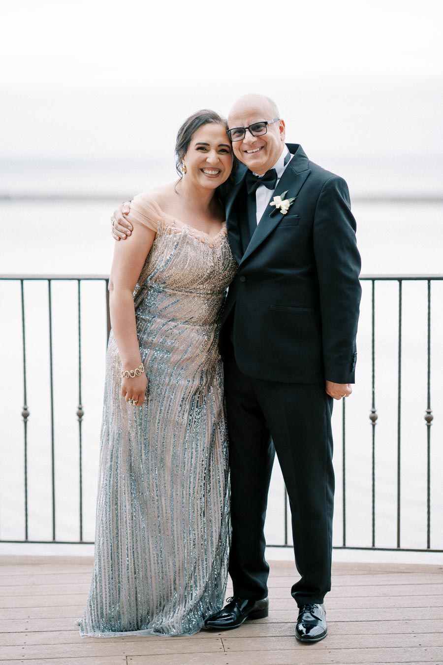Elegant couple posing together at an outdoor event, woman in a sparkling dress and man in a tuxedo, smiling against a waterfront backdrop.