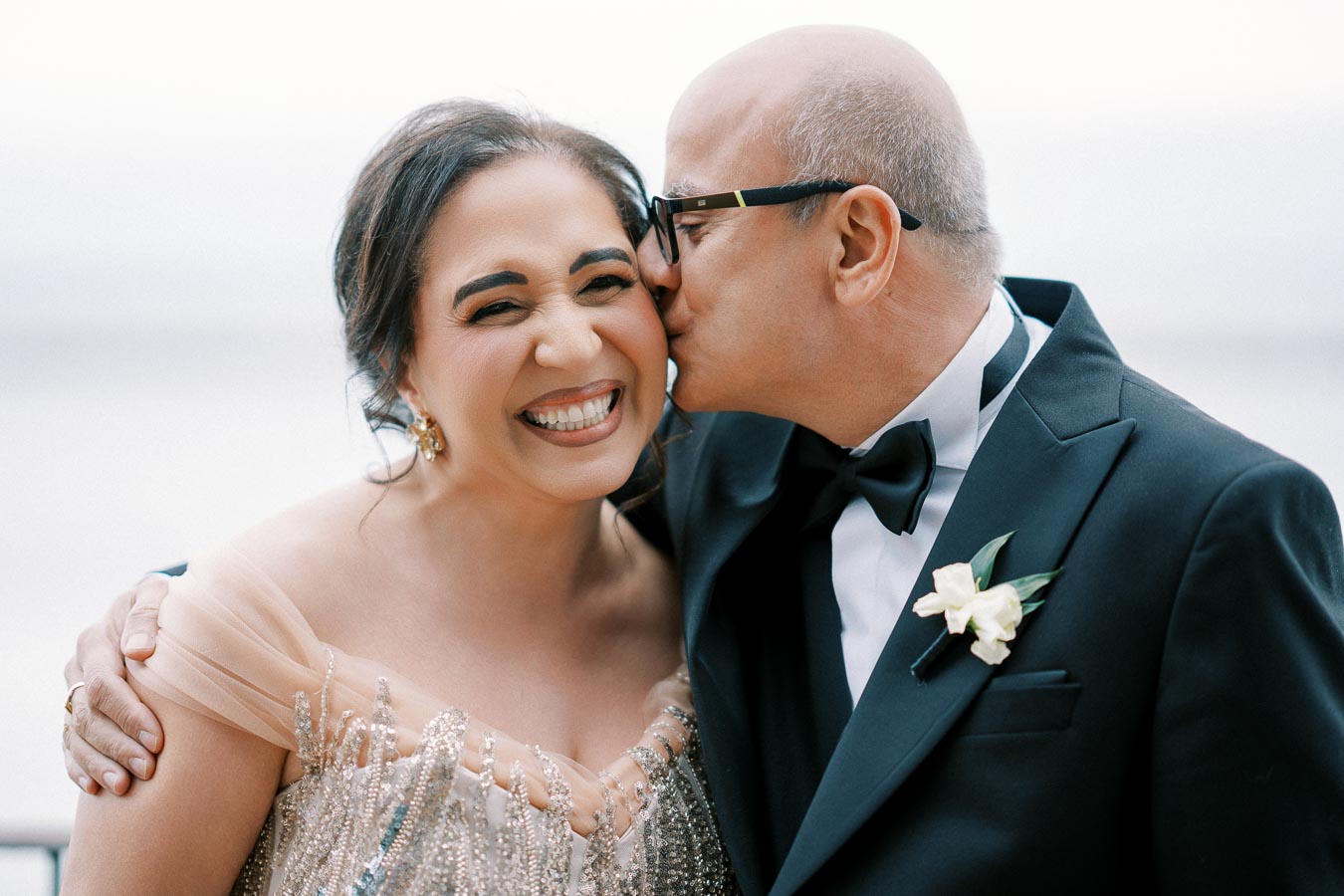 Elderly couple at a formal event, with the man in a tuxedo kissing the smiling woman wearing an elegant gown, evoking warmth and joy.