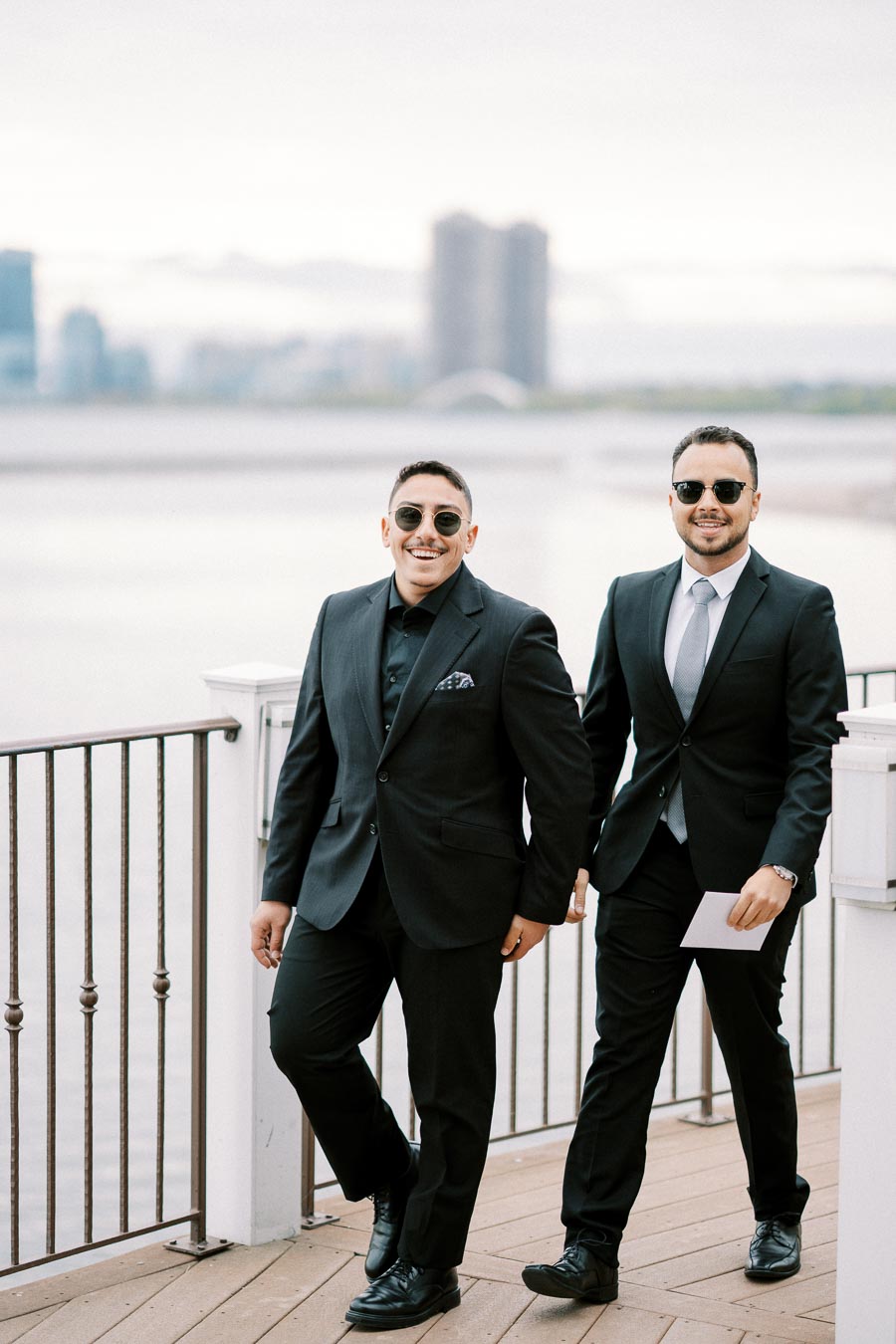 Two men in suits walking on a waterfront deck, smiling and wearing sunglasses, with a city skyline in the background.