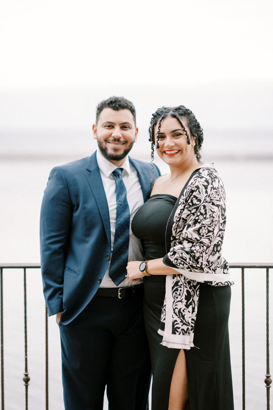 Elegantly dressed couple smiling and standing by a waterfront railing, with a scenic background, wearing formal attire – a man in a suit and a woman in a black dress and patterned shawl.