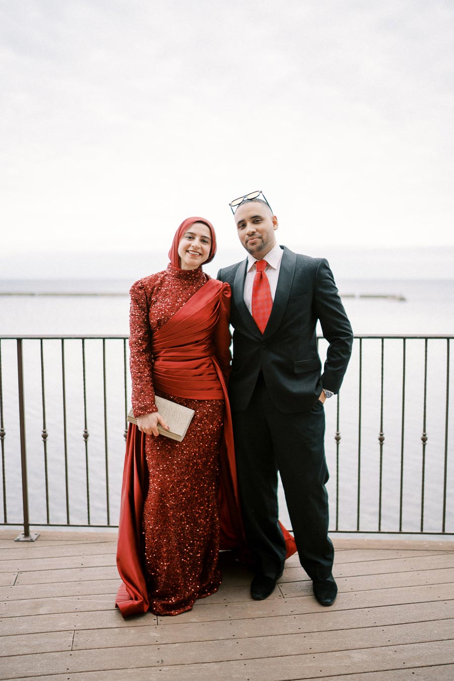 A woman in a sparkling red gown and hijab stands next to a man in a black suit with a red tie, posing on a deck overlooking a calm body of water.
