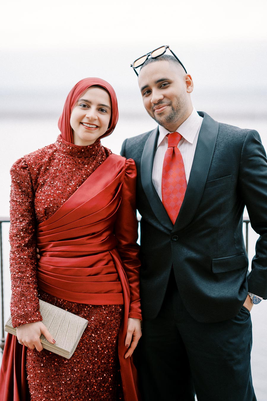 Certainly! Here is a suitable alt text you can use for this image:Smiling, well-dressed couple posing in elegant attire; the woman in a sparkling red gown with a matching hijab, and the man in a black suit with a red tie, standing against a blurred background.