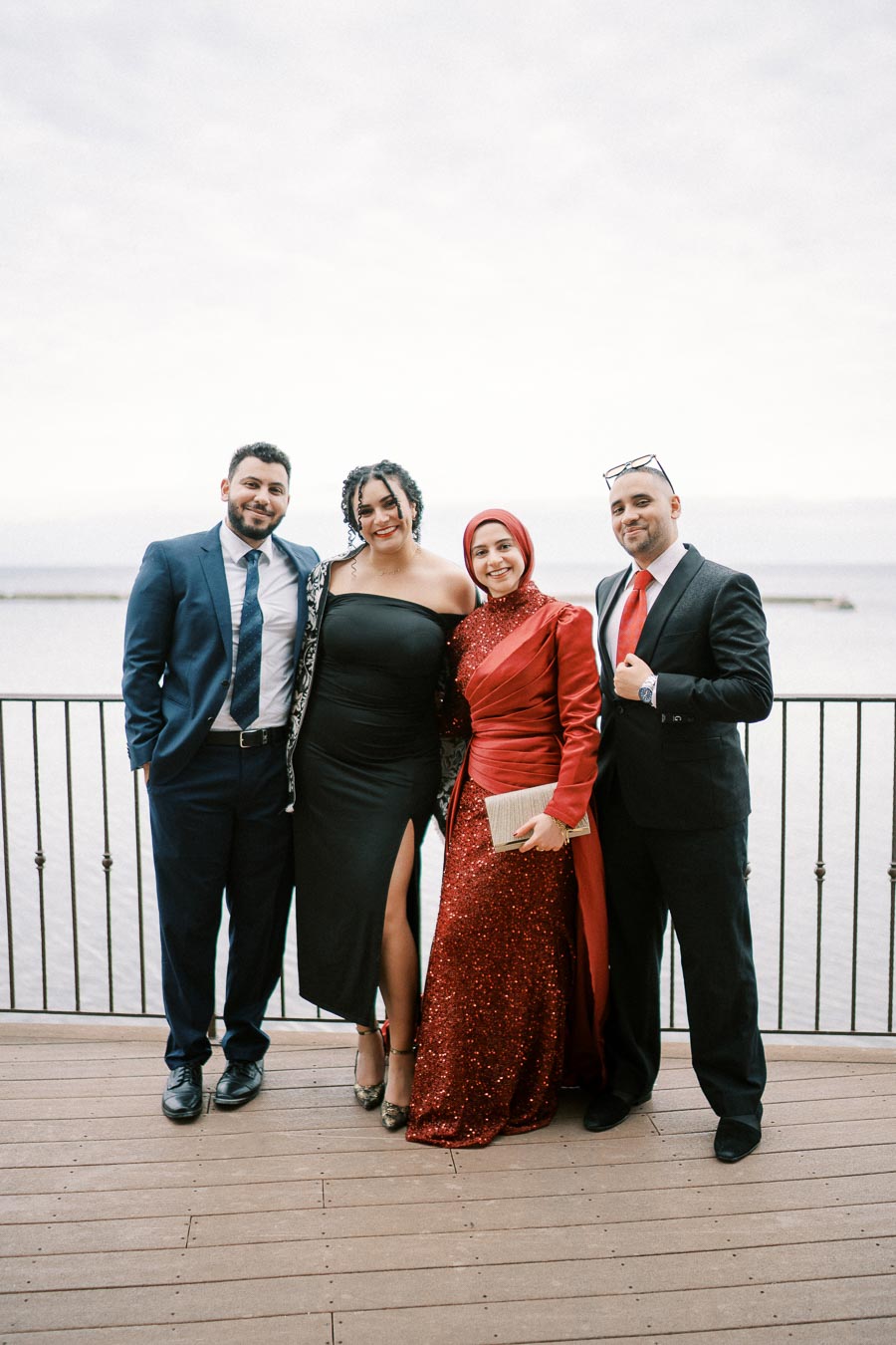 Four elegantly dressed individuals smiling and posing on a wooden deck with a scenic waterfront backdrop. The group features formal attire, including suits and dresses, ideal for a special occasion or event by the water.
