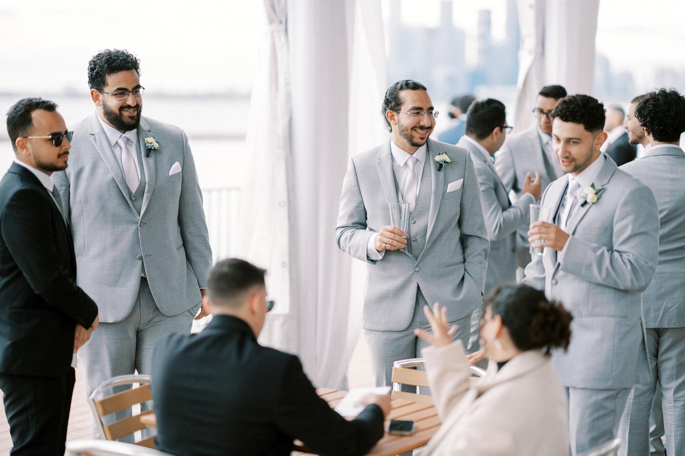 Group of groomsmen in light gray suits and ties conversing joyfully at a wedding reception, with a city skyline in the background. Exchanging laughter and drinks, they create a warm and celebratory atmosphere.