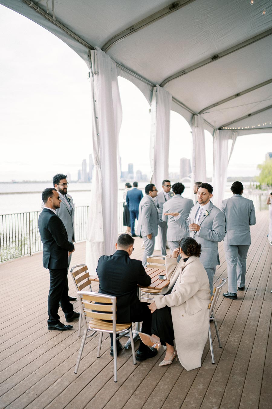 People socializing under a stylish canopy at an outdoor event with a city skyline and waterfront view in the background, featuring attendees in formal attire and business suits.