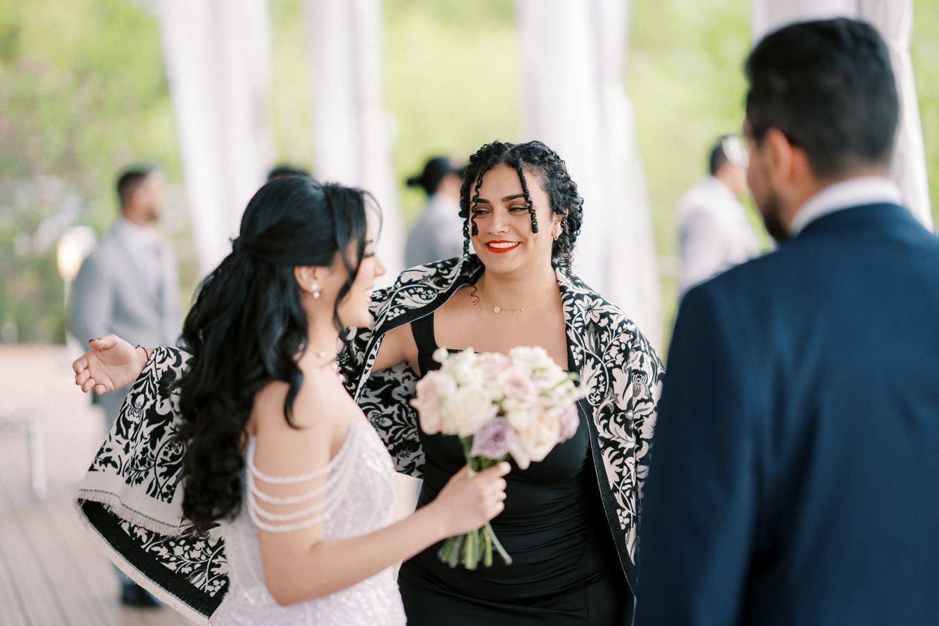 Woman in elegant dress holding a floral bouquet, smiling and greeting guests at an outdoor event.