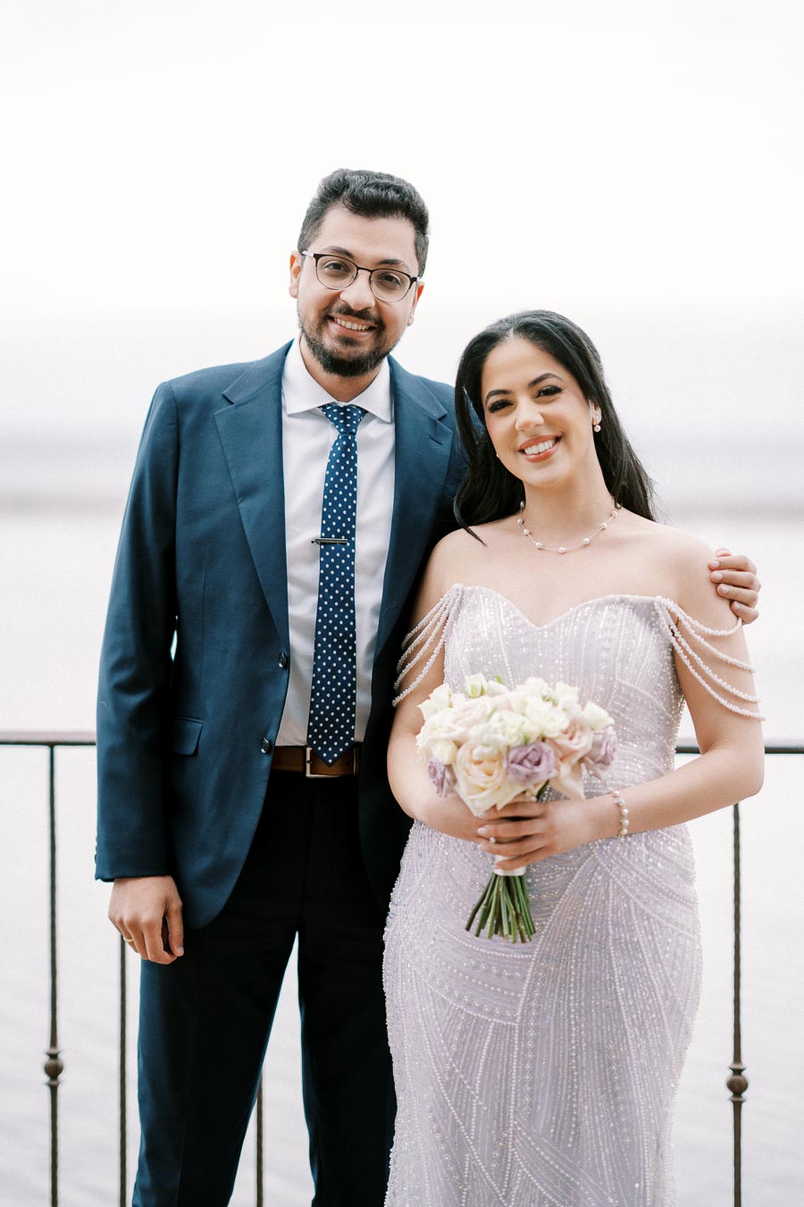 Smiling couple in formal attire by a waterfront; woman in an elegant beaded gown holding a bouquet of pastel roses, man in a blue suit and polka dot tie.