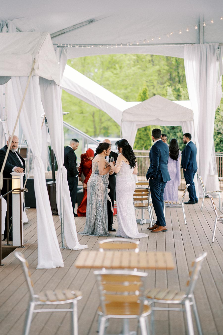 Outdoor wedding reception under a white tent, featuring elegantly dressed guests mingling and socializing on a wooden deck with lush greenery in the background.