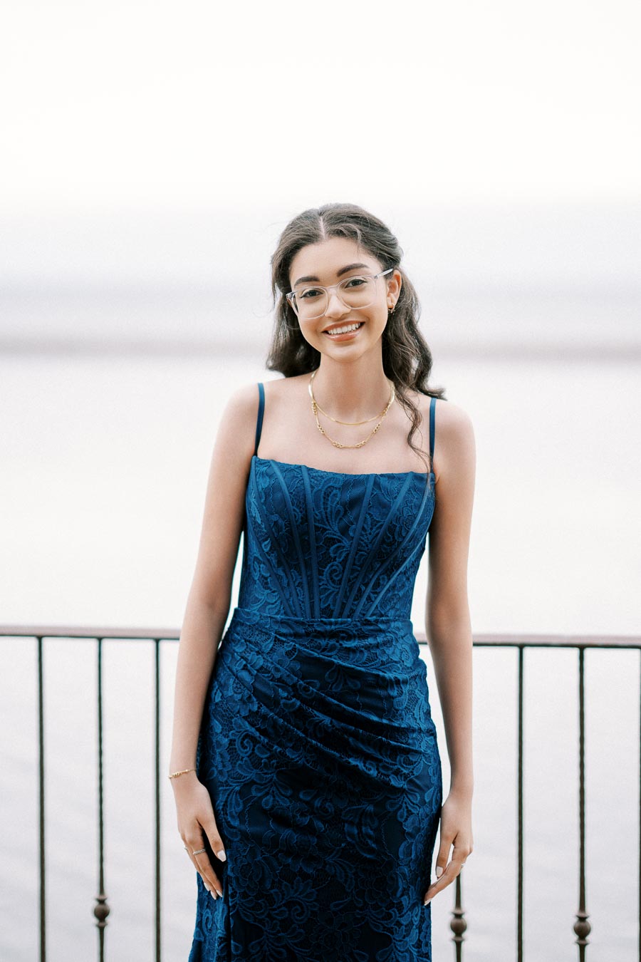 A young woman in a blue, intricately patterned formal dress stands smiling on a balcony with a soft, blurred background of serene water and sky, exuding elegance and confidence.