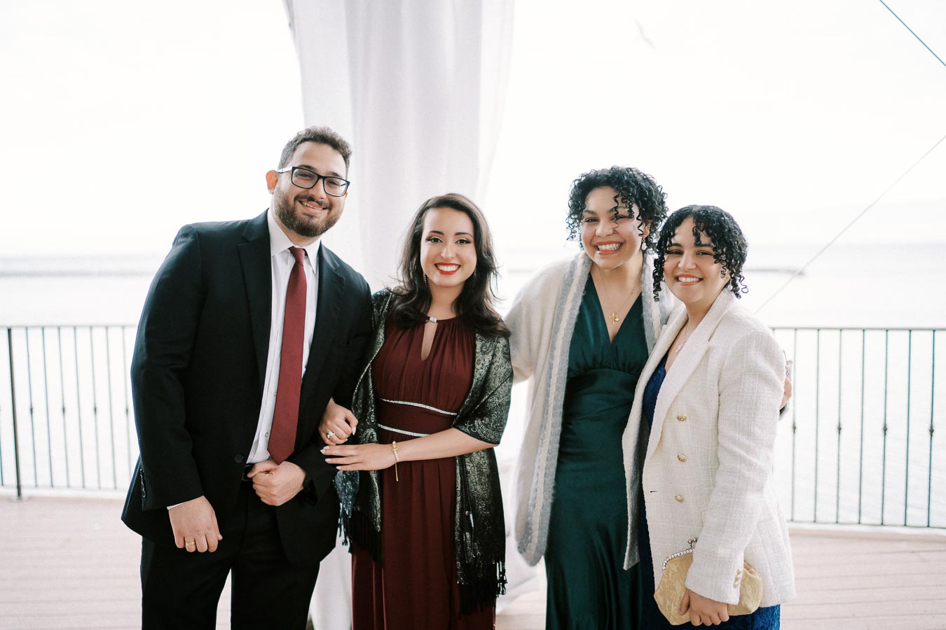 A group of four smiling people dressed in formal attire, standing together on a balcony with a view of the ocean in the background.