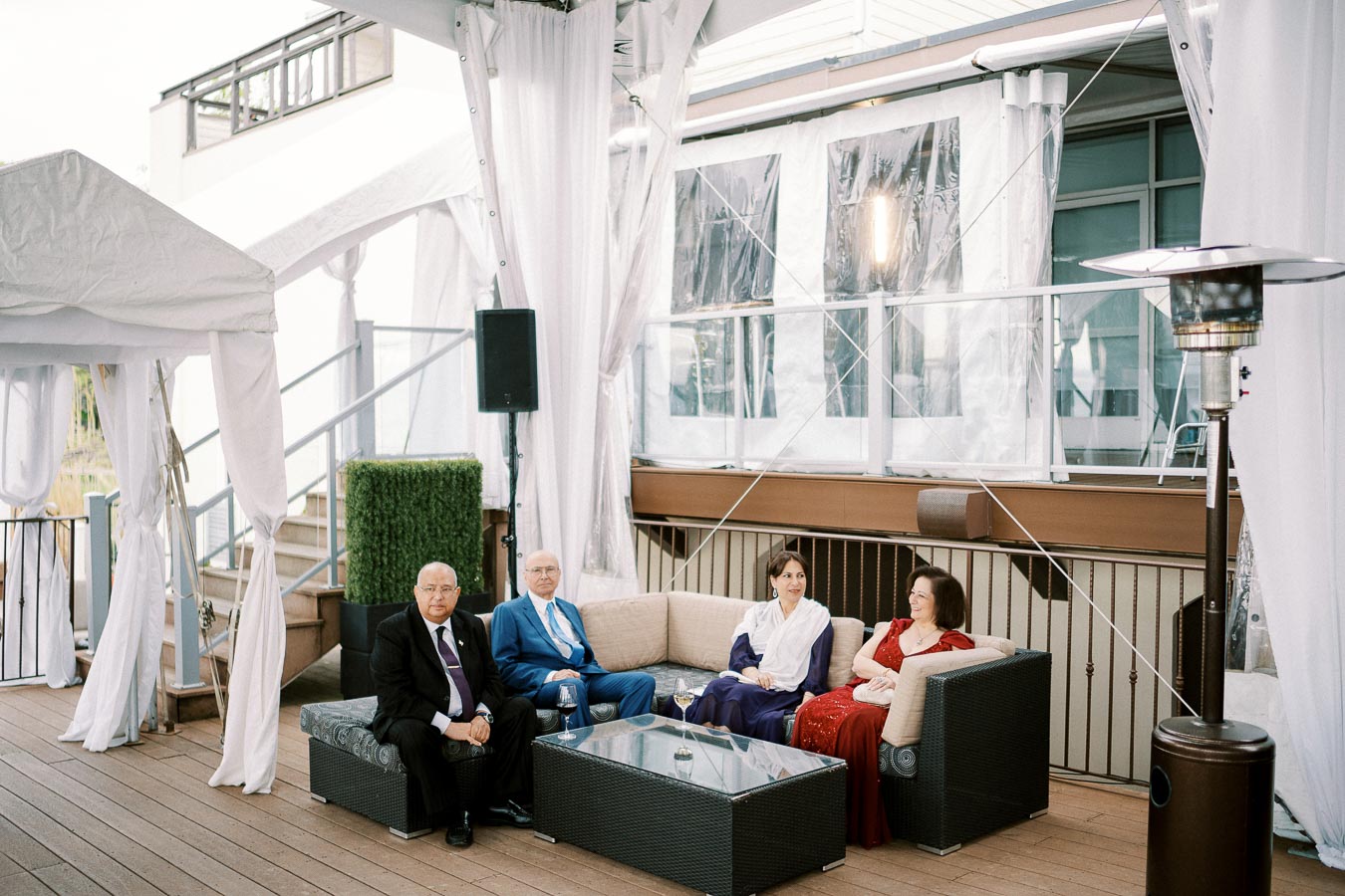 Four elegantly dressed individuals sitting on a patio sofa under a white tent, with a modern building in the background, creating a sophisticated outdoor gathering setting.