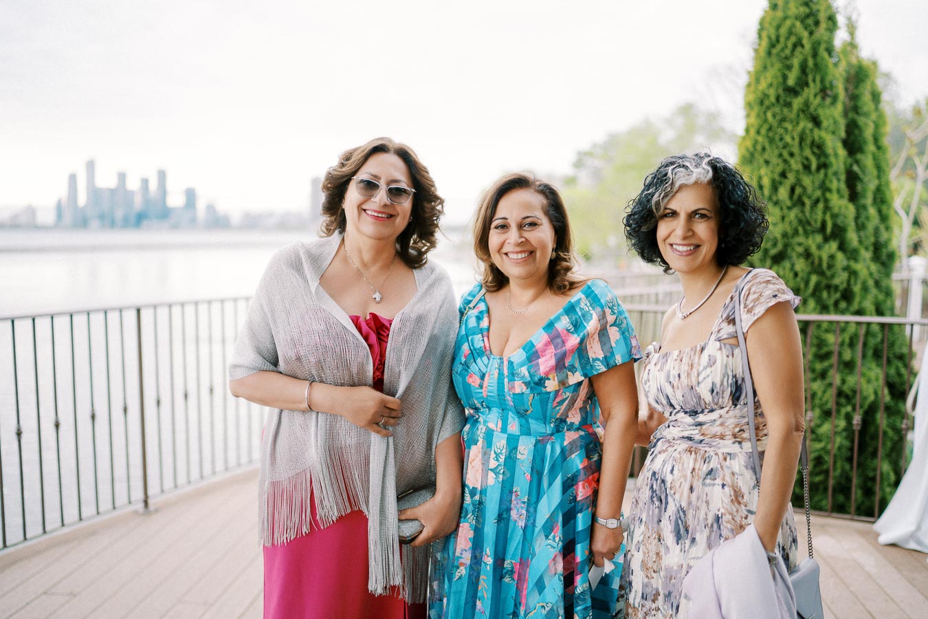 Three women pose on a waterfront balcony with a city skyline in the background, wearing colorful dresses and smiling.