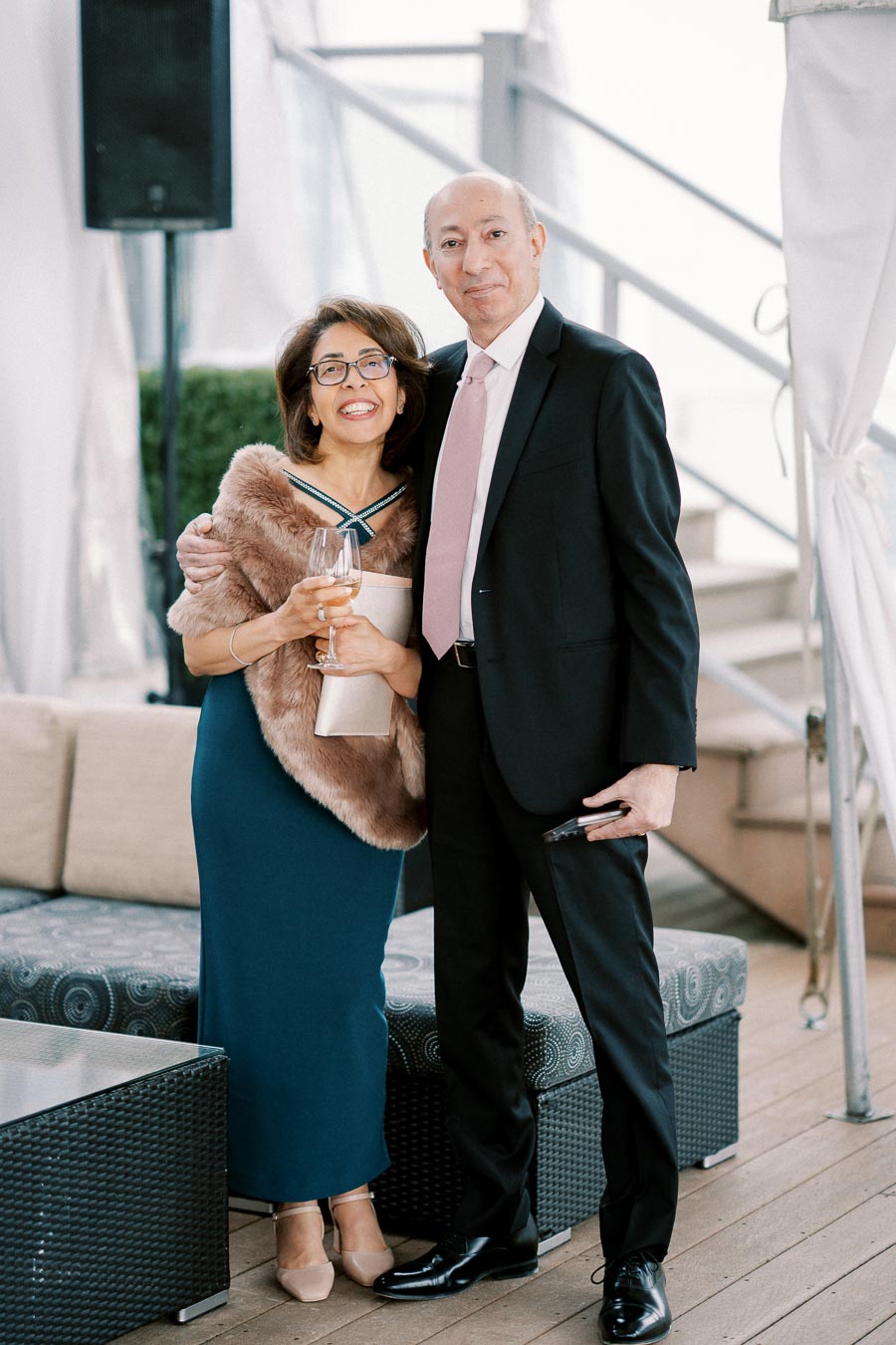 A smiling couple elegantly dressed for a formal event, standing together on a stylish patio. The woman is holding a wine glass and a clutch, while the man is in a suit holding a phone, with a modern, sophisticated backdrop.