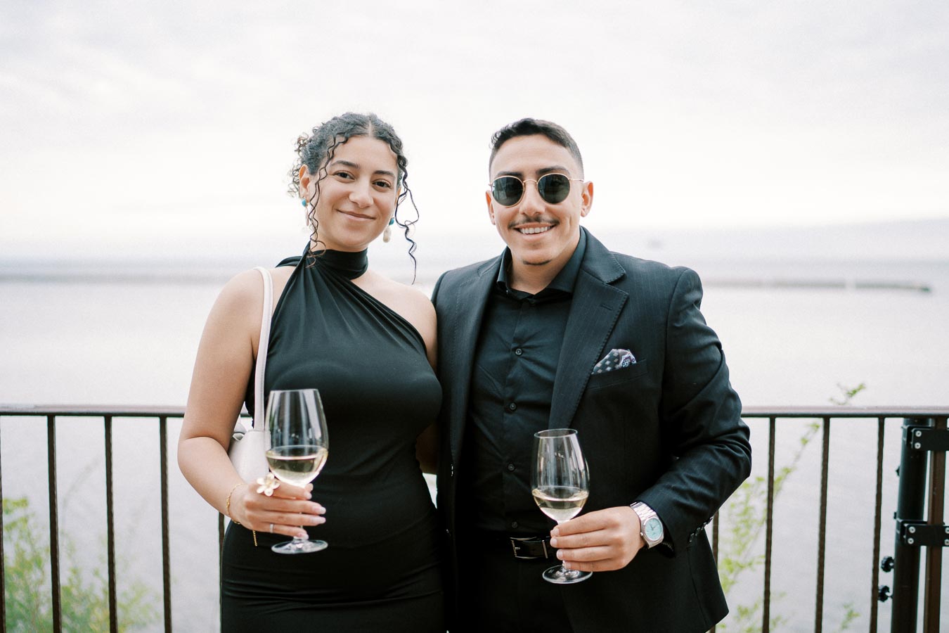 A couple elegantly dressed in black outfits holding wine glasses, smiling at the camera with a scenic waterfront background.