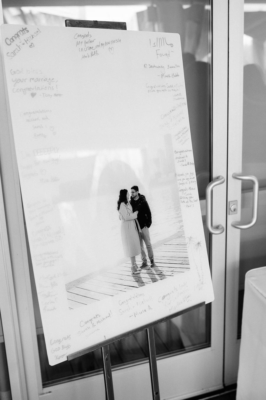 A black and white image of a large poster board featuring a romantic couple standing together on a wooden pier. The board is filled with handwritten congratulatory messages for their engagement. The setting appears to be a celebratory event, possibly an engagement party, with glass doors in the background.
