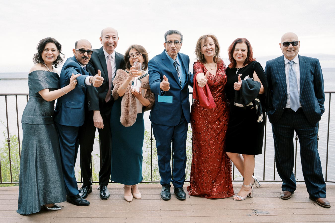 A group of elegantly dressed people standing together on a wooden deck, smiling and giving thumbs up, with a scenic waterfront view in the background.