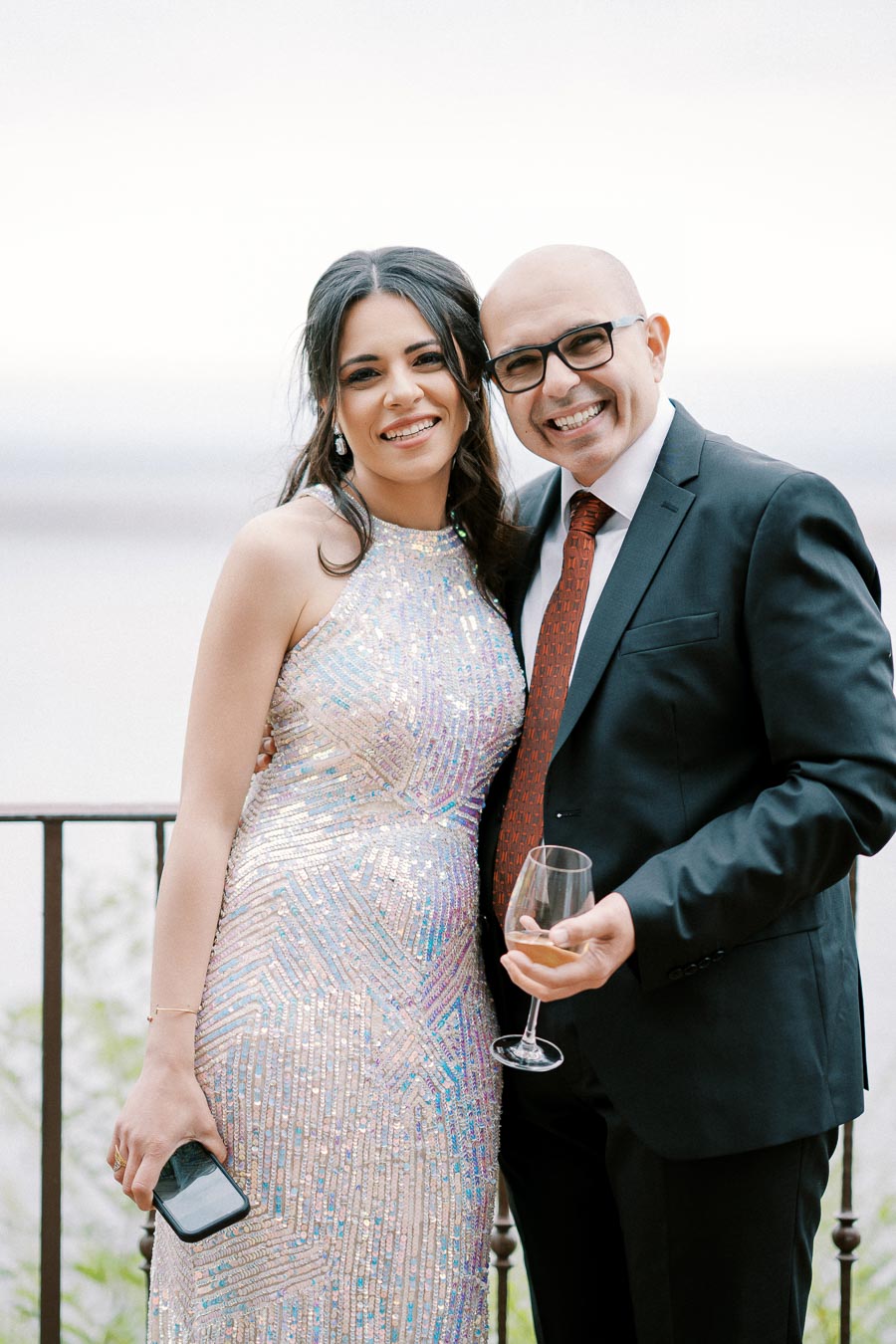 Elegant couple smiling at formal event, woman in sequin dress holding phone, man in suit with wine glass against outdoor backdrop.