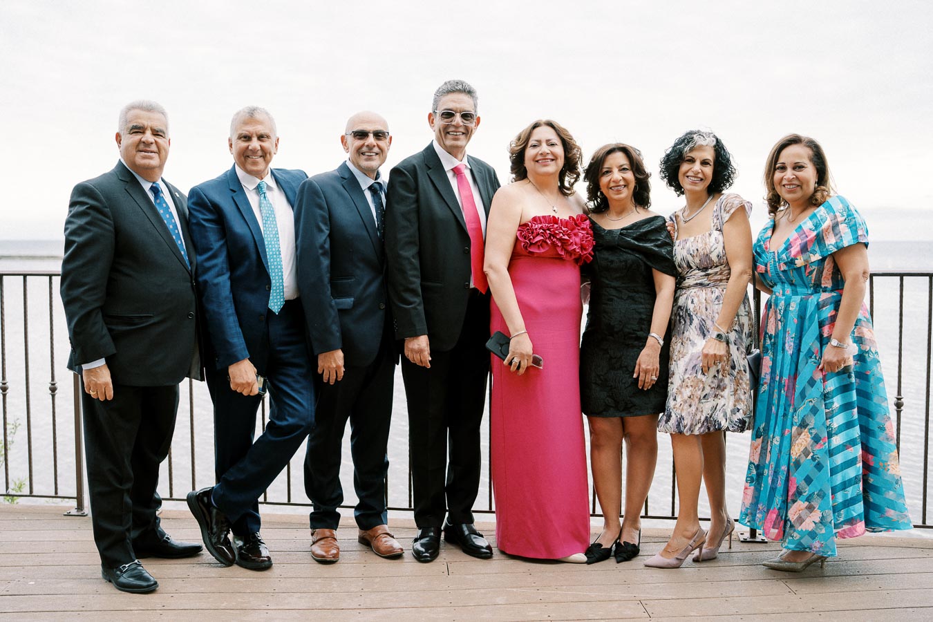 A group of well-dressed individuals smiling on a deck overlooking water, men in suits and women in elegant dresses, suggesting a formal event or celebration.