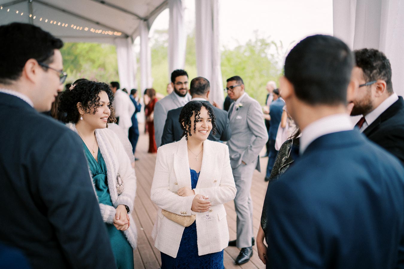 Group of elegantly dressed people socializing at an outdoor event under a tent, with a focus on a woman in a white blazer smiling and holding a drink.