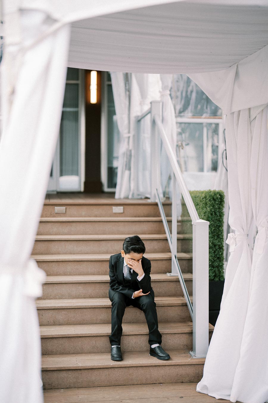 Young boy in a suit sitting on stairs, covering his face, under a white canopy.