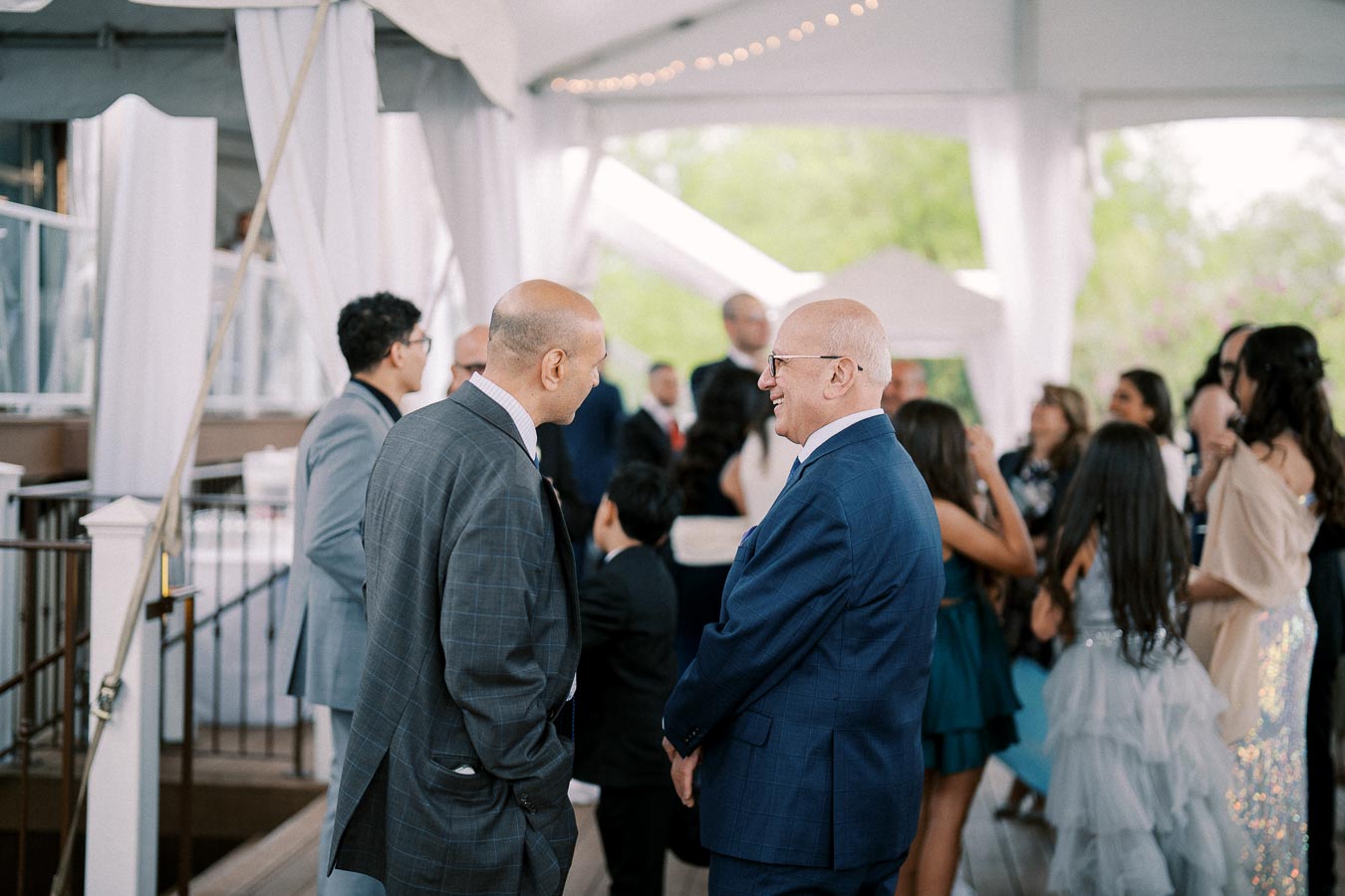 A group of well-dressed people socializing at an indoor event, featuring two men in suits engaged in conversation, surrounded by a lively gathering under a white canopy.